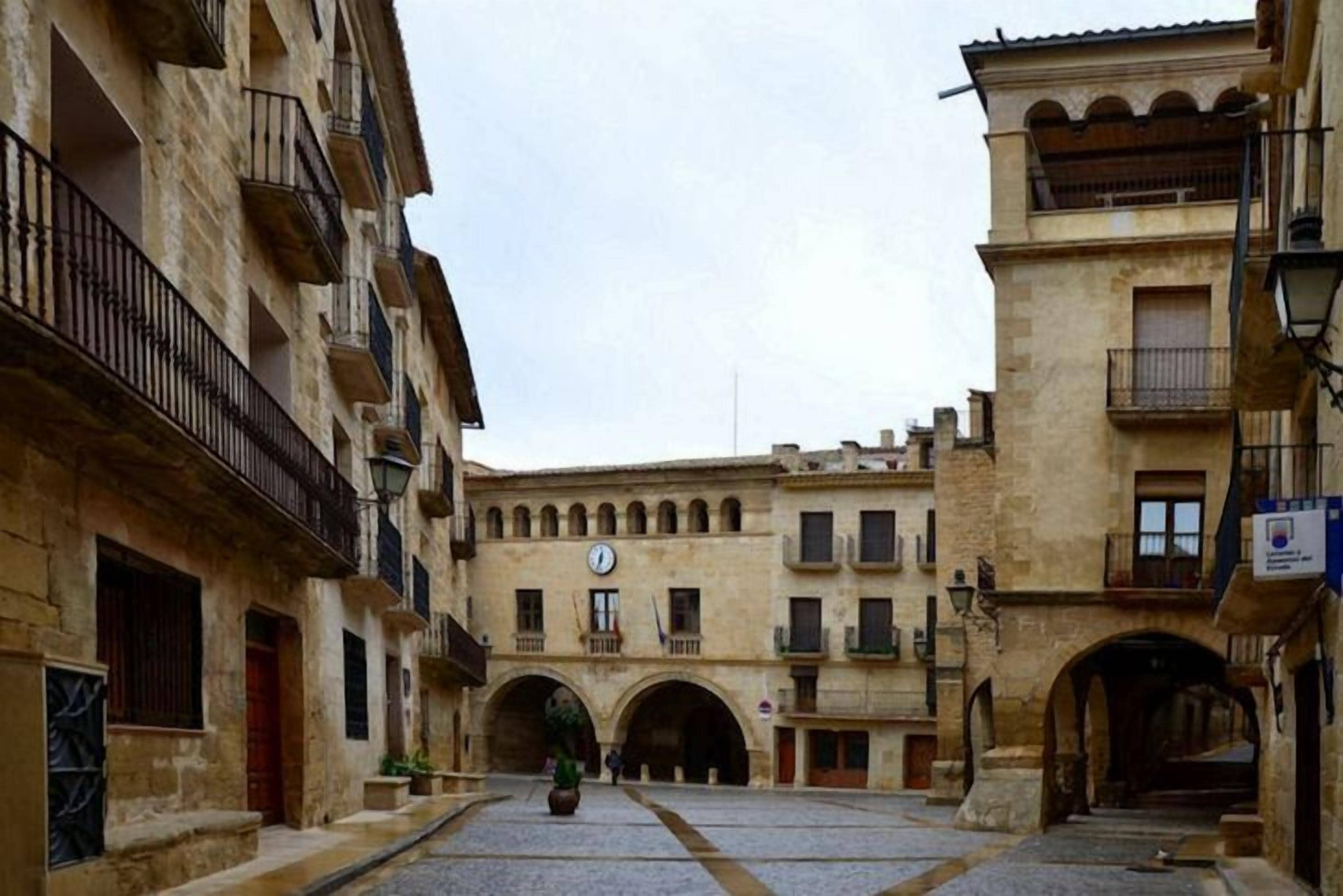 Una plaza histórica con edificios de piedra, paseos con arcos y una torre del reloj en el centro.