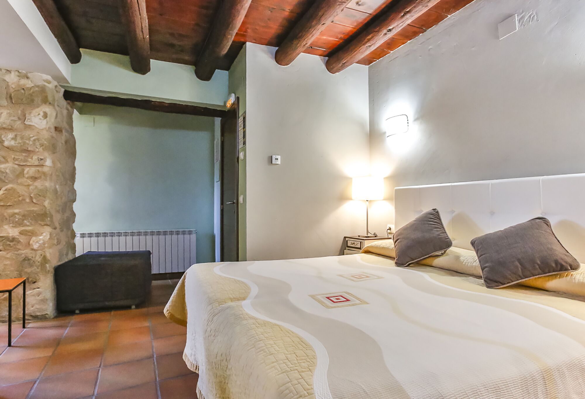 Ein Schlafzimmer mit einem Bett, einem Nachttisch, einer Lampe, einer Steinwand und einer Holzbalkendecke.
