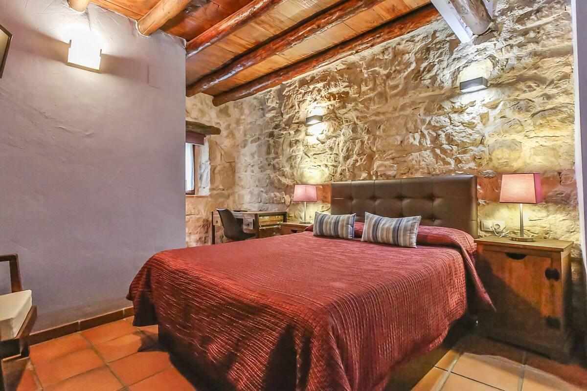A bedroom with a stone wall, a wooden ceiling, a bed with a red bedspread, two bedside tables with lamps, and a chair.