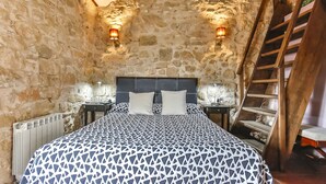 A bedroom with a stone wall, a bed with patterned bedding, two bedside lamps, and a wooden ladder. - La Grancha (La Fresneda)