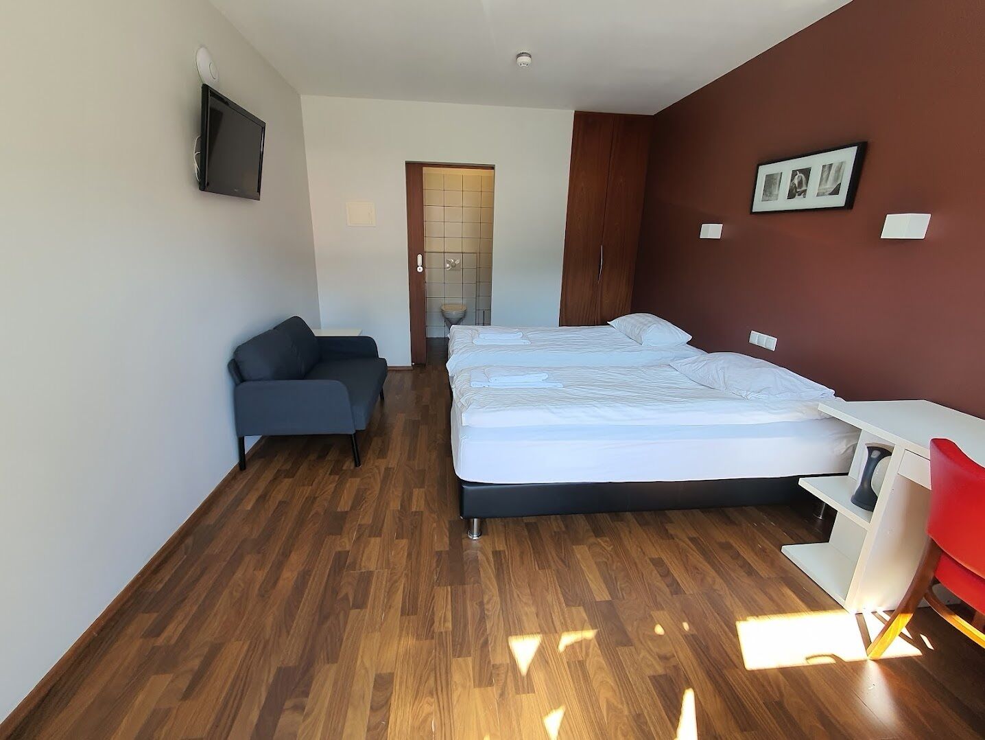 Een moderne hotelkamer met een houten vloer, een bed met witte lakens, een grijze bank, een bureau en een aan de muur bevestigde televisie.