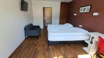 Een moderne hotelkamer met een houten vloer, een bed met witte lakens, een grijze bank, een bureau en een aan de muur bevestigde televisie.