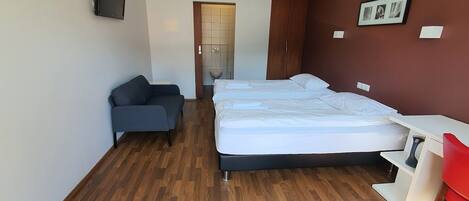 A modern hotel room with a wooden floor, a bed with white linens, a grey sofa, a desk, and a TV mounted on the wall.
