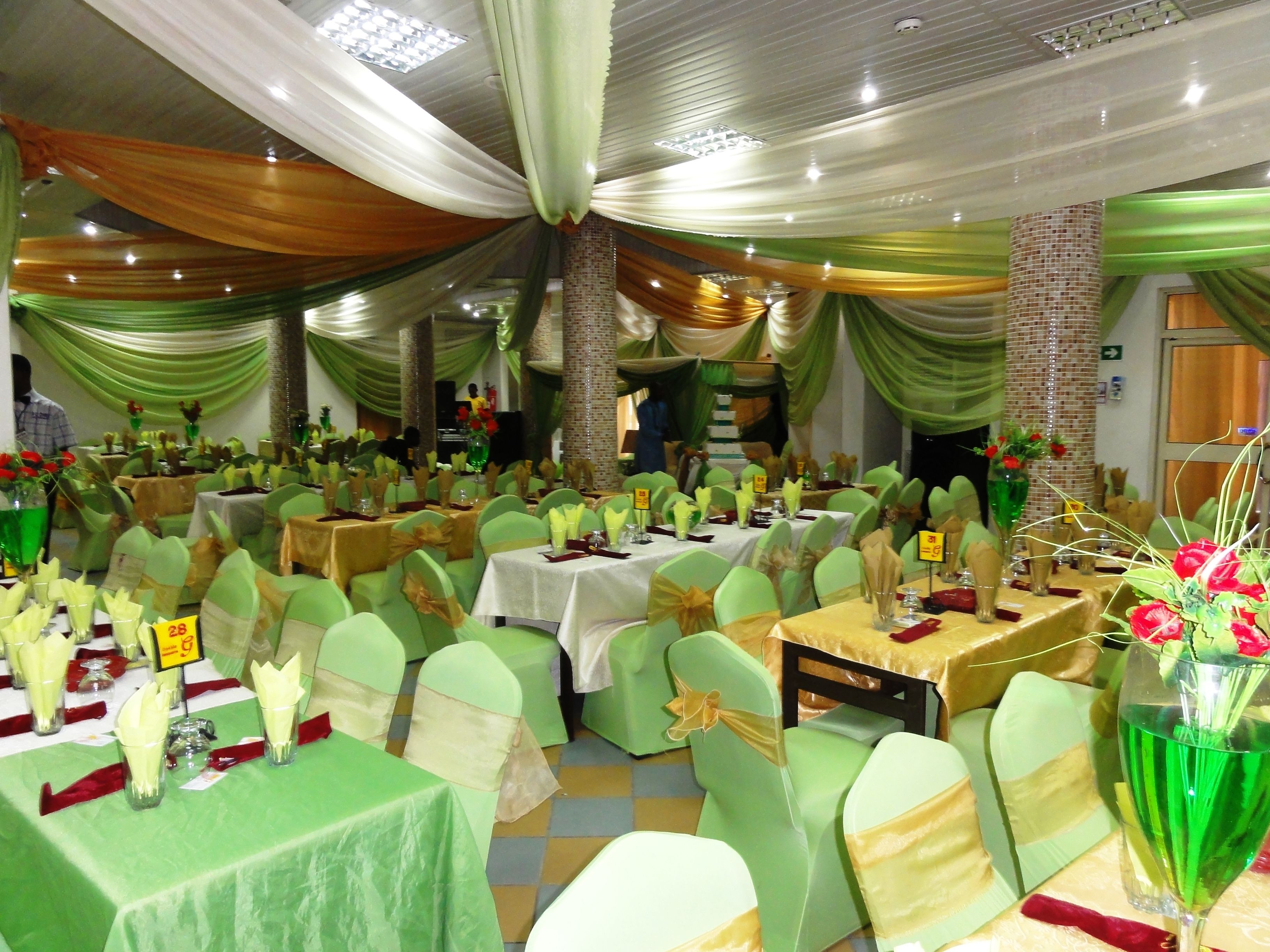 Un salón de banquetes con mesas dispuestas para un evento formal, con decoración en verde y dorado, y una columna de ladrillo en el centro.