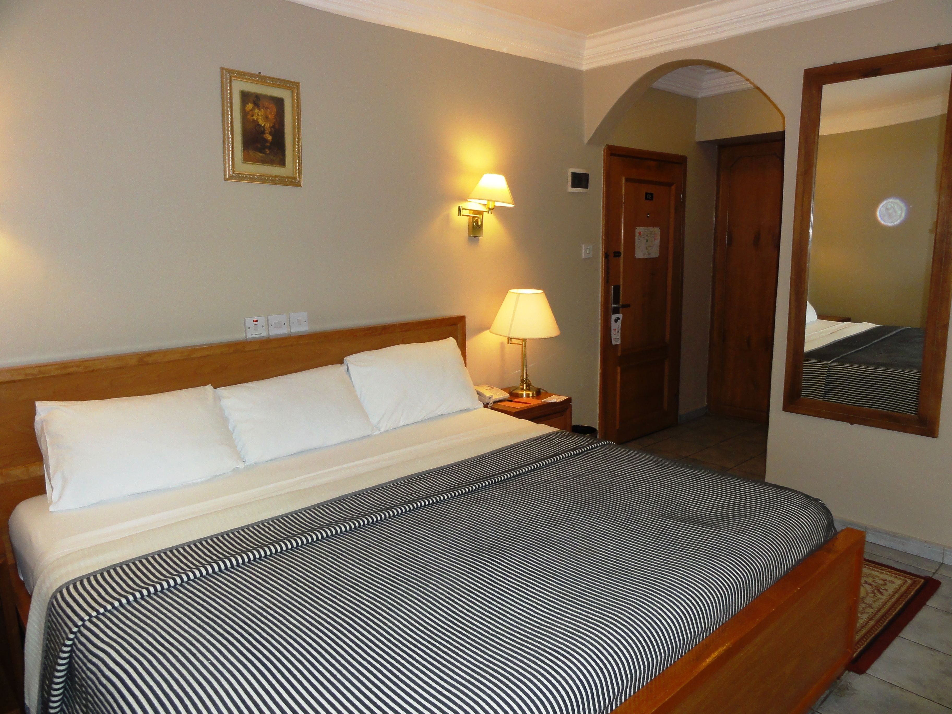 A hotel room with a bed, a nightstand with a lamp, a wall-mounted light, a door, and a mirror.