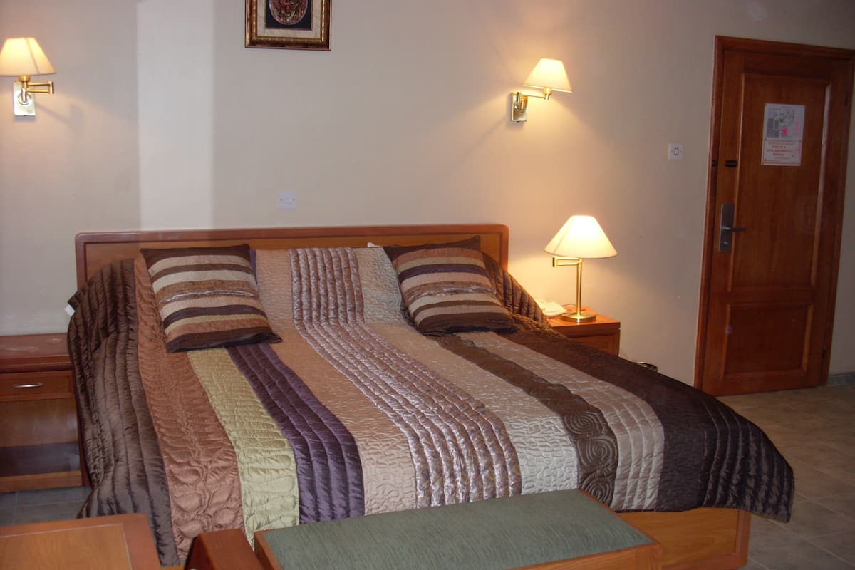 Ein ordentlich bezogenes Bett mit gestreiftem Bettbezug und Kissen, zwei Nachttischlampen, eine Holzbank und eine Tür mit einem Schild.