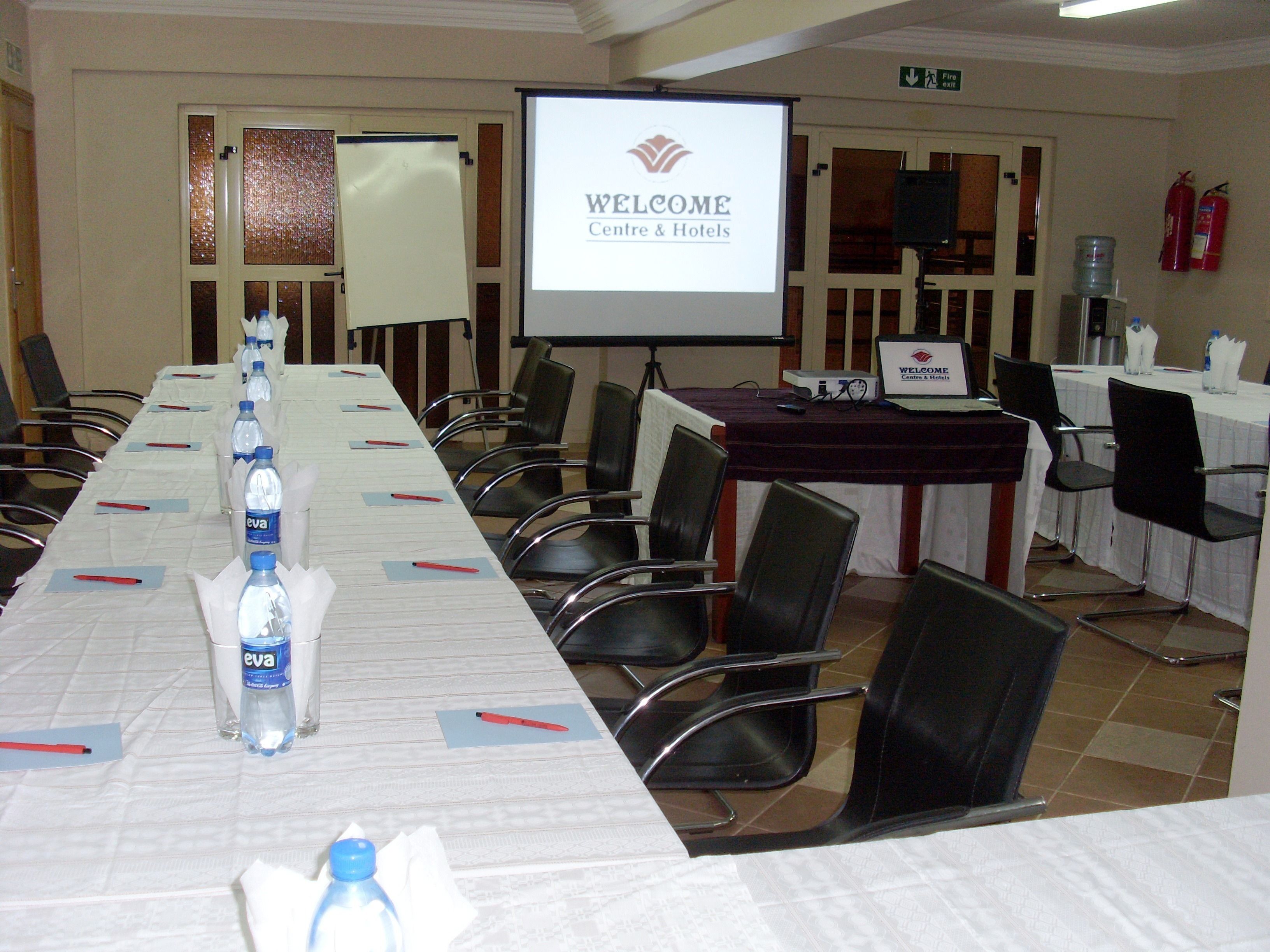 A conference room with a long table, chairs, a projector screen displaying "WELCOME Centre & Hotels", and a water dispenser.