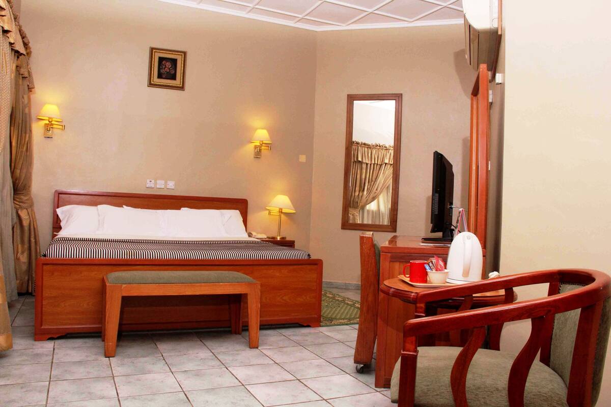 Ein Hotelzimmer mit einem hölzernen Bett, Nachttischlampen, einem Schreibtisch mit Stuhl, einem Fernseher und einem Spiegel.