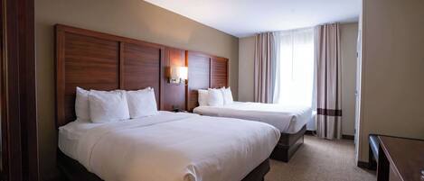 A hotel room with two beds, a wooden headboard, a desk, and a large window with curtains.