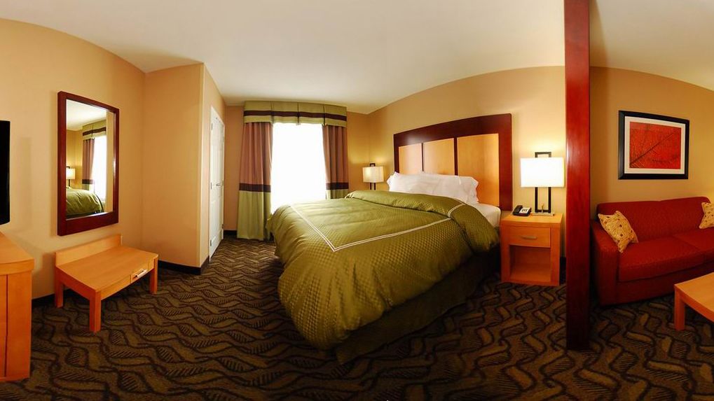 A hotel room with a large bed, a desk, a chair, a red sofa, a nightstand with a lamp, and a mirror on the wall.
