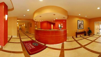 Zona de recepción de un hotel con un mostrador curvo, una alfombra roja con el logotipo de 'Comfort Suites' y un pasillo con suelo de baldosas.