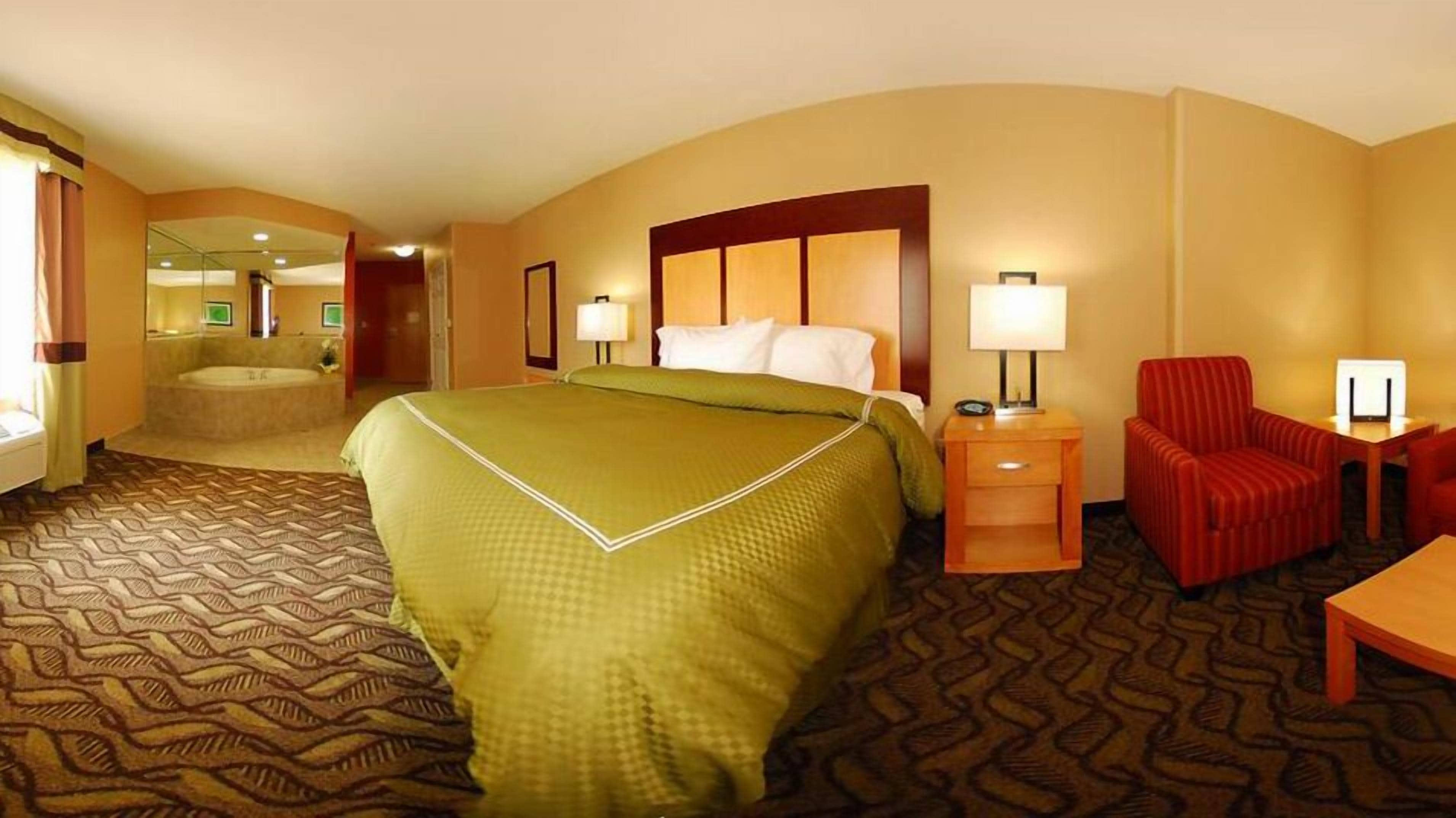 Habitación de hotel con una cama grande, un escritorio, una silla, una mesita de noche, una lámpara y un baño con bañera y retrete.