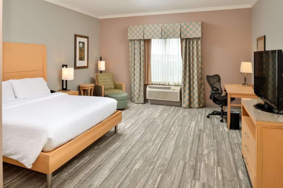 A hotel room with a bed, two chairs, a desk with a television, an air conditioning unit, and a window with curtains.