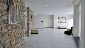 A spacious lobby with a stone wall, a large abstract painting, a seating area, and a door in the background. - Hilton Garden Inn Denton (Denton)
