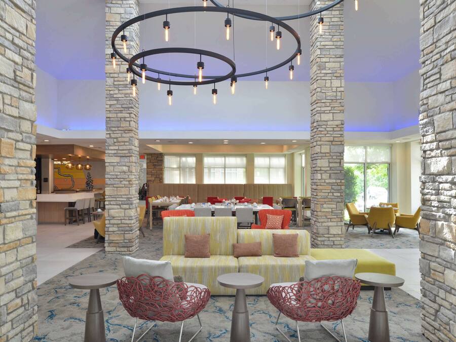 A lobby with stone pillars, a central seating area with sofas and chairs, a dining area in the background, and a modern chandelier.