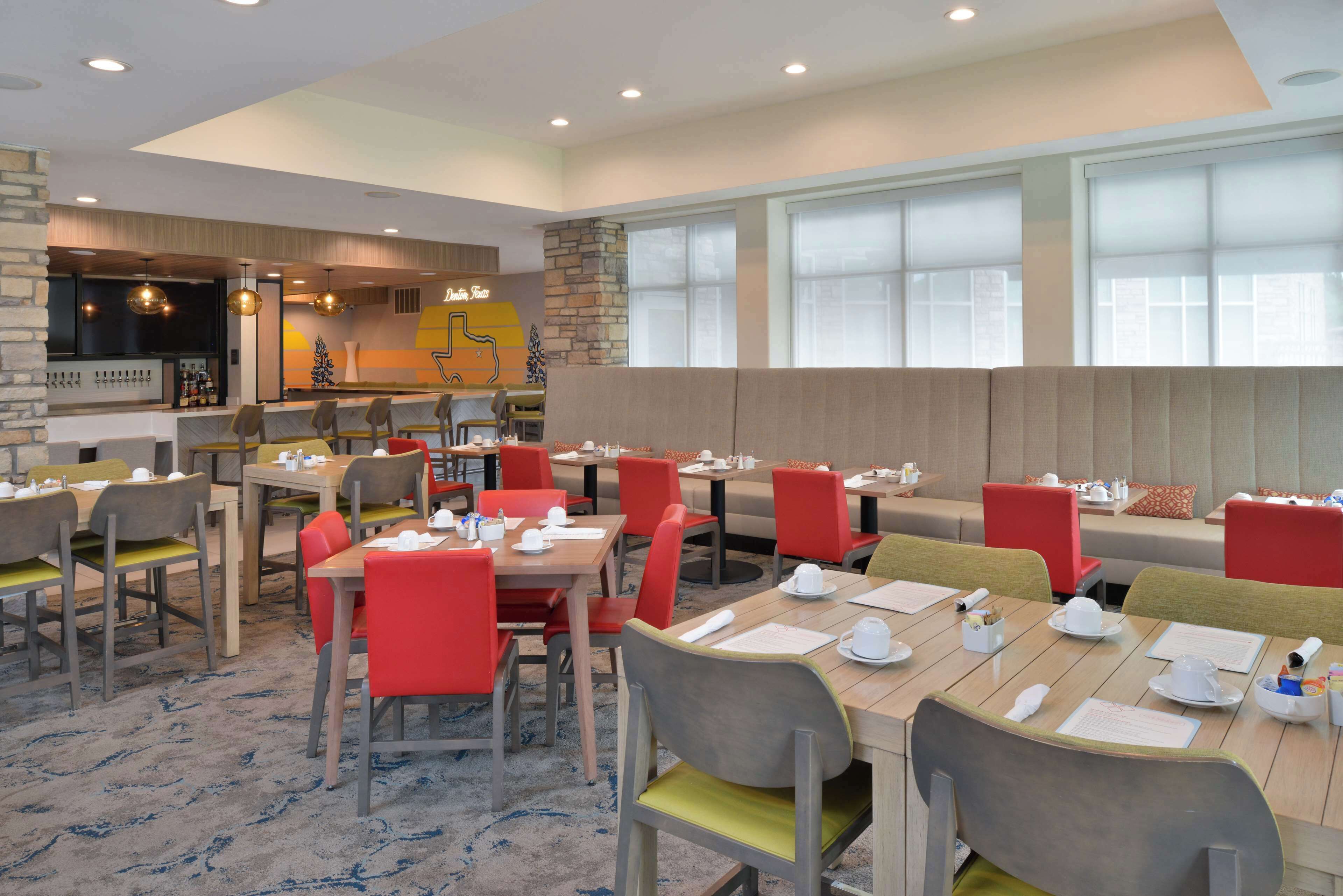 Un restaurante con varias mesas y sillas, una zona de bar y ventanas amplias que dejan entrar luz natural.