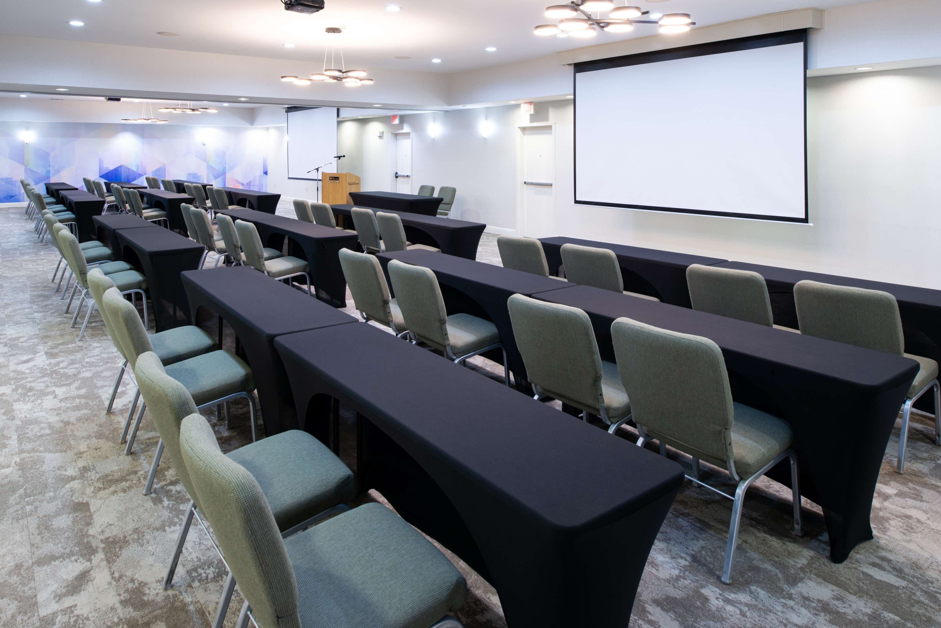 Un salón de conferencias con filas de mesas y sillas, una pantalla de proyector y suelo alfombrado.
