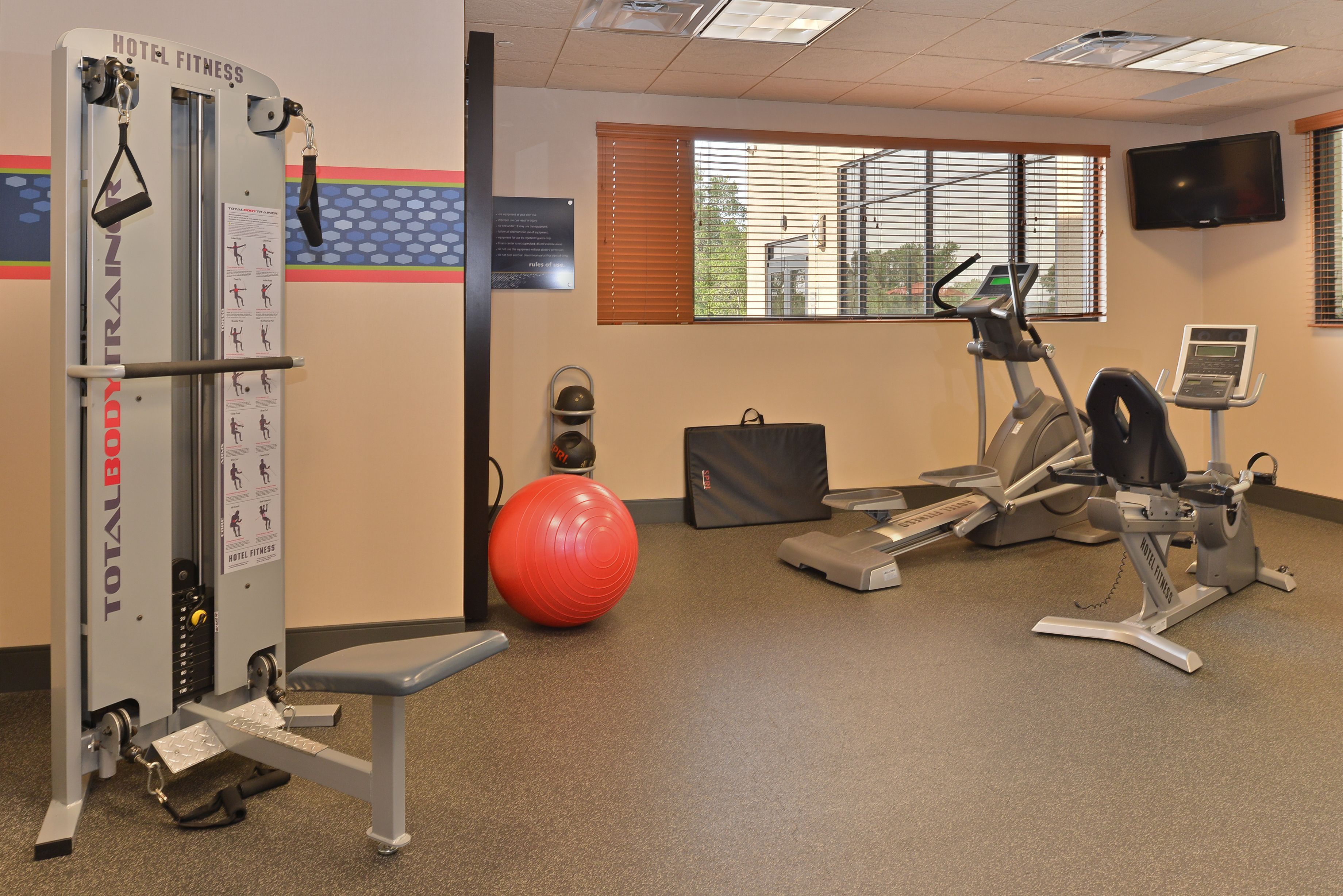 Un gimnasio de hotel con varios equipos de ejercicio, incluyendo una máquina de entrenamiento integral, una elíptica y una pelota de ejercicios roja.