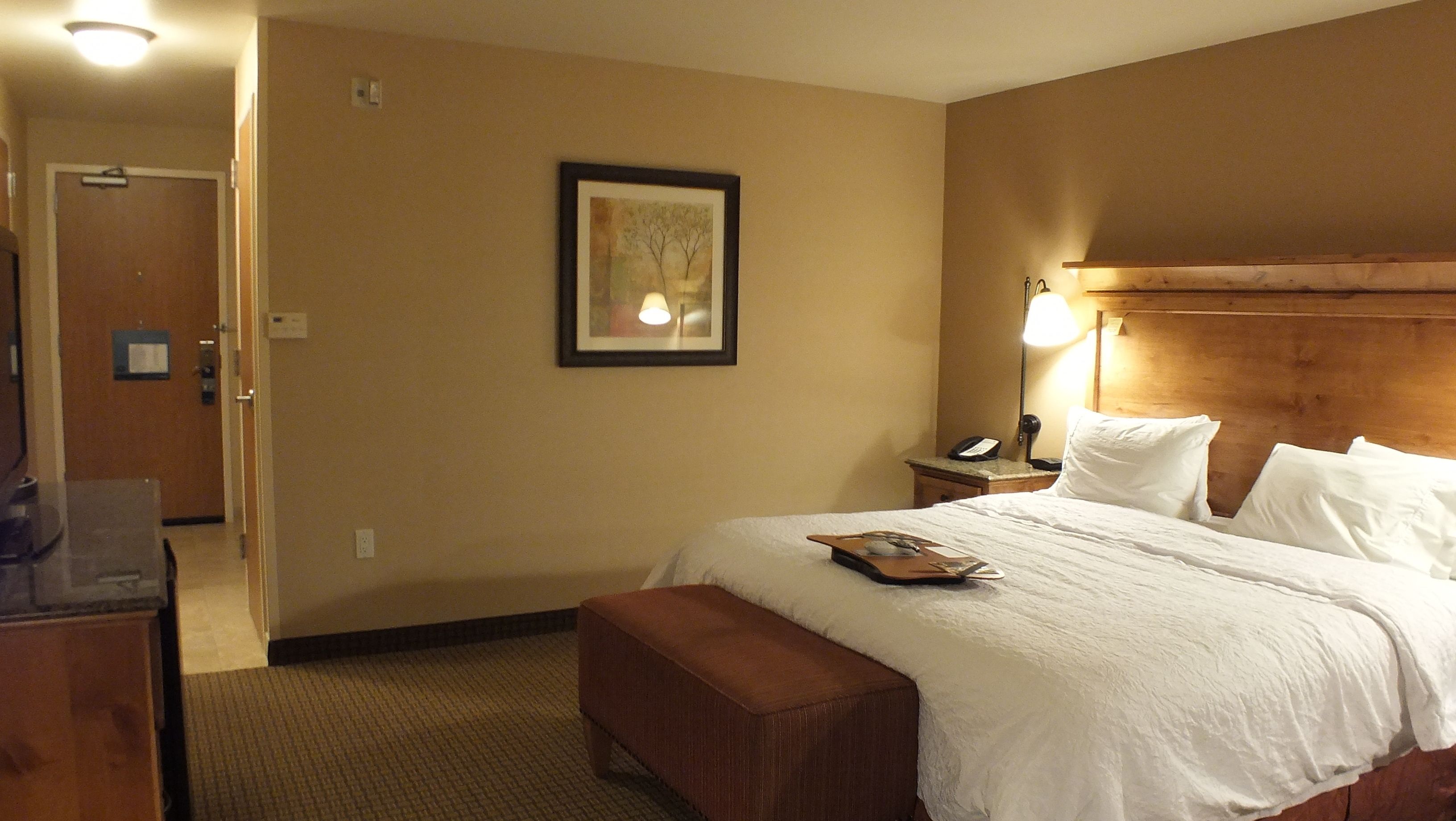 A hotel room with a large bed, a bedside table with a lamp, a framed picture on the wall, and a wooden headboard.