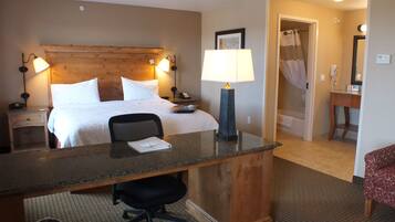 A hotel room with a bed, bedside lamps, a desk with a chair, a bathroom with a shower, and a mirror.