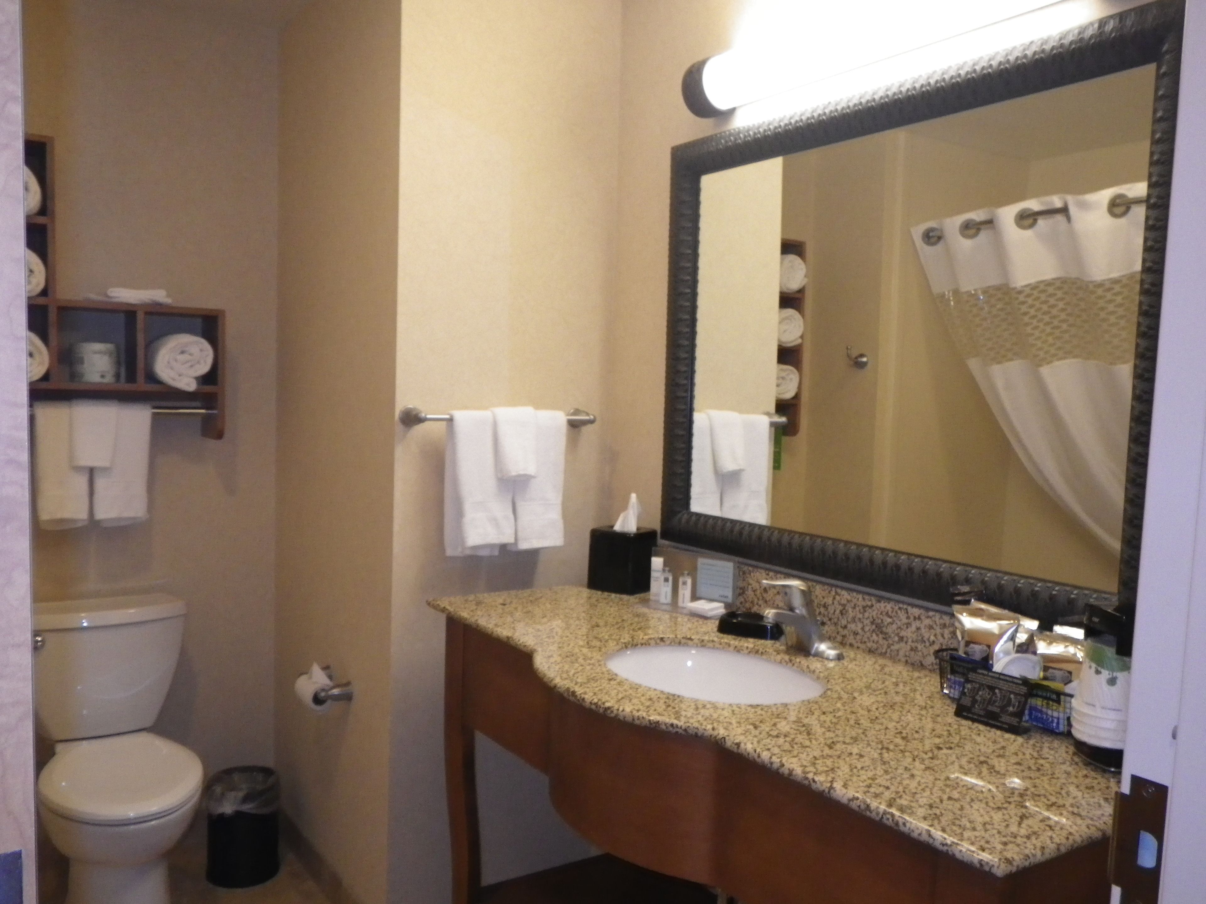 A bathroom with a toilet, sink, and mirror. There are towels hanging on a rack and a shelf with toiletries.