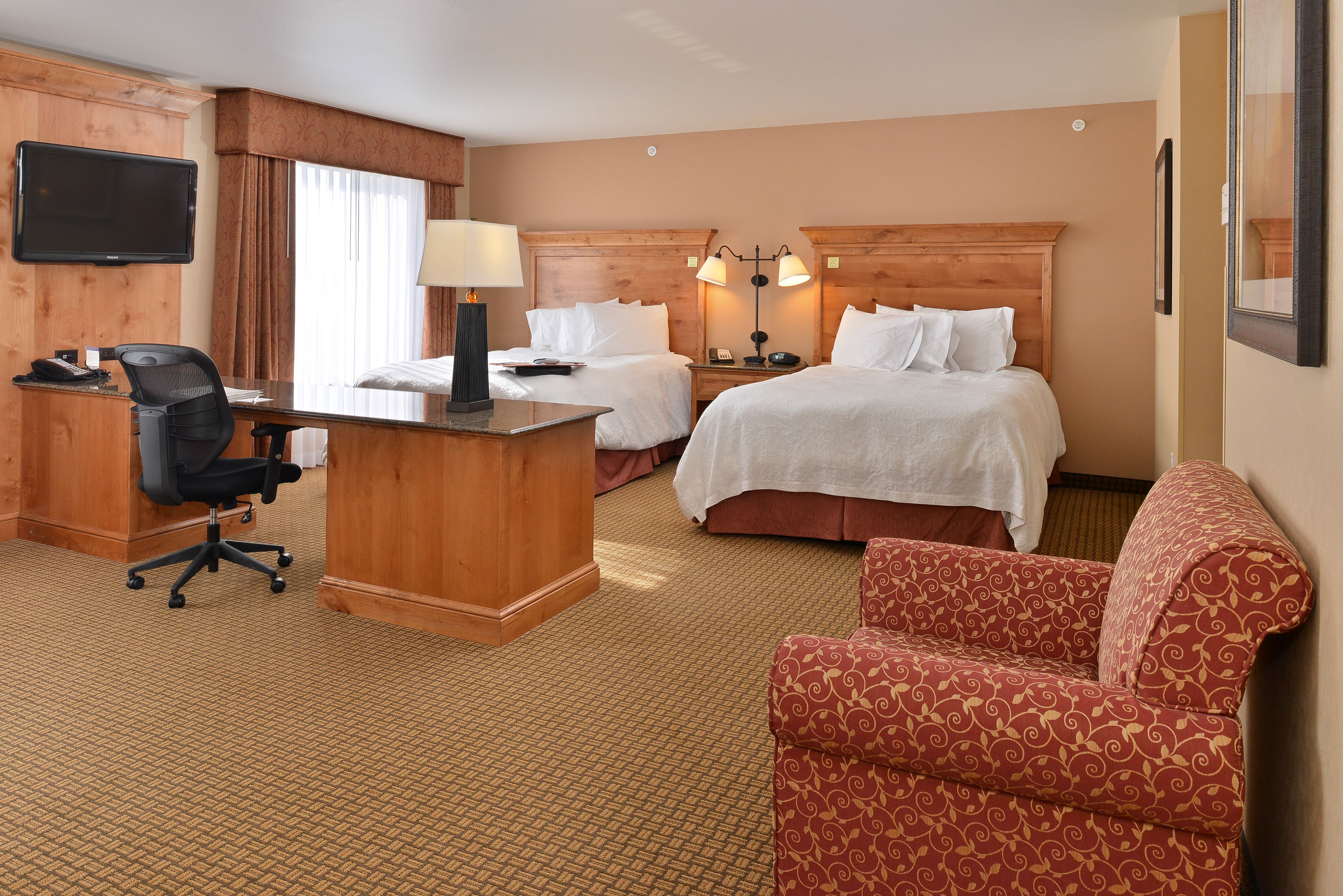 A hotel room with two beds, a desk with a chair, a TV mounted on the wall, and a red patterned armchair.