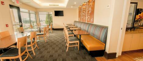 A hotel dining area with multiple tables and chairs, a long bench, and a television on the wall.