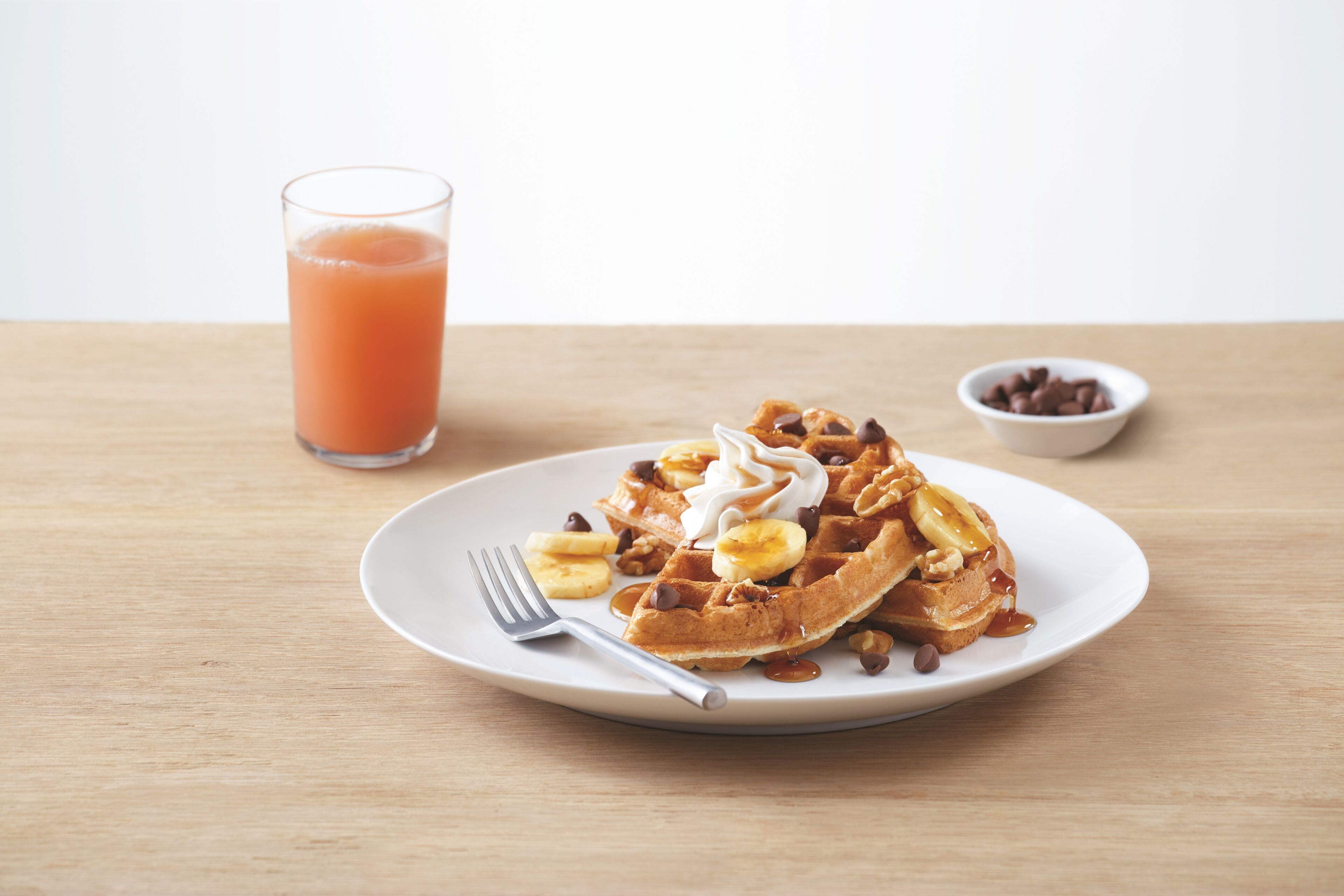 Uma pilha de waffles com bananas, gotas de chocolate e chantilly, um copo de sumo de laranja e uma taça com gotas de chocolate sobre uma mesa de madeira.