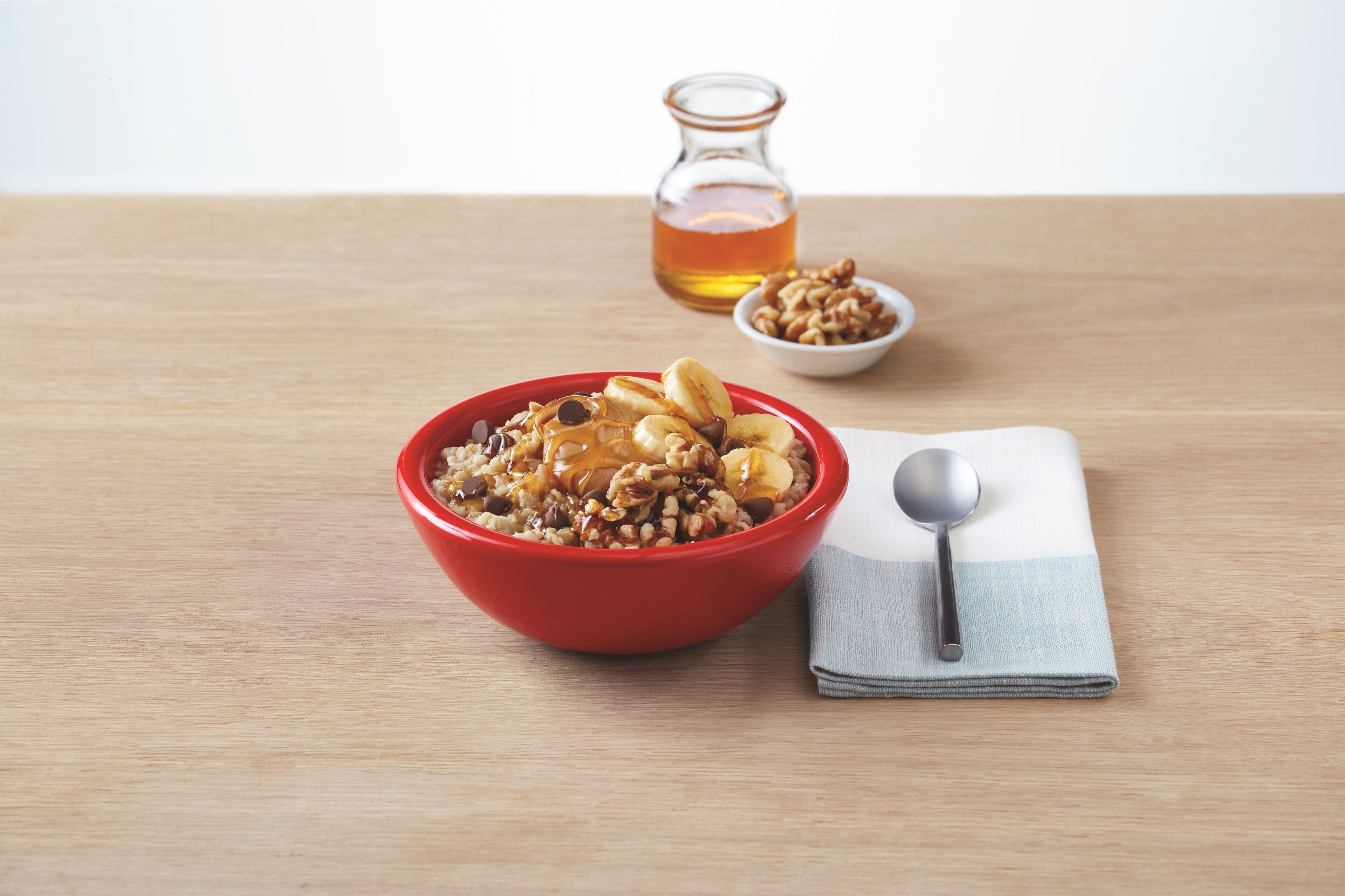 A bowl of cereal with a spoon and a bottle of syrup on a wooden table.