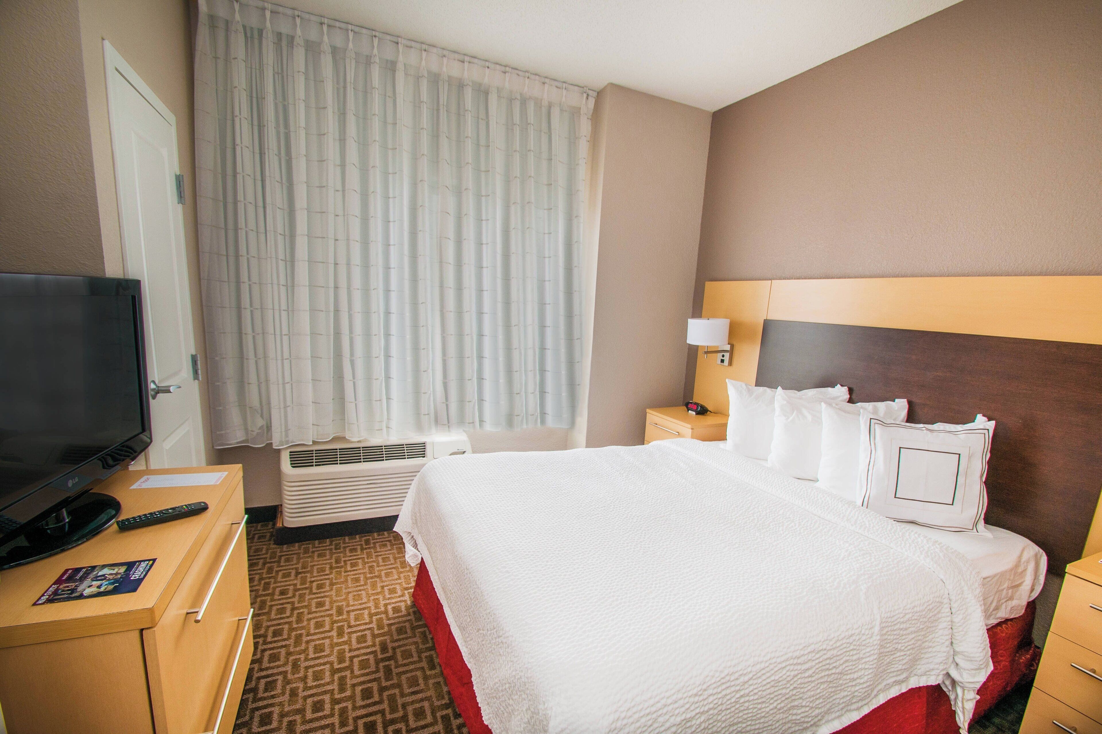 Ein Hotelzimmer mit einem Bett, einem Fernseher, einem Schreibtisch mit Fernbedienung und einem Fenster mit Vorhängen.