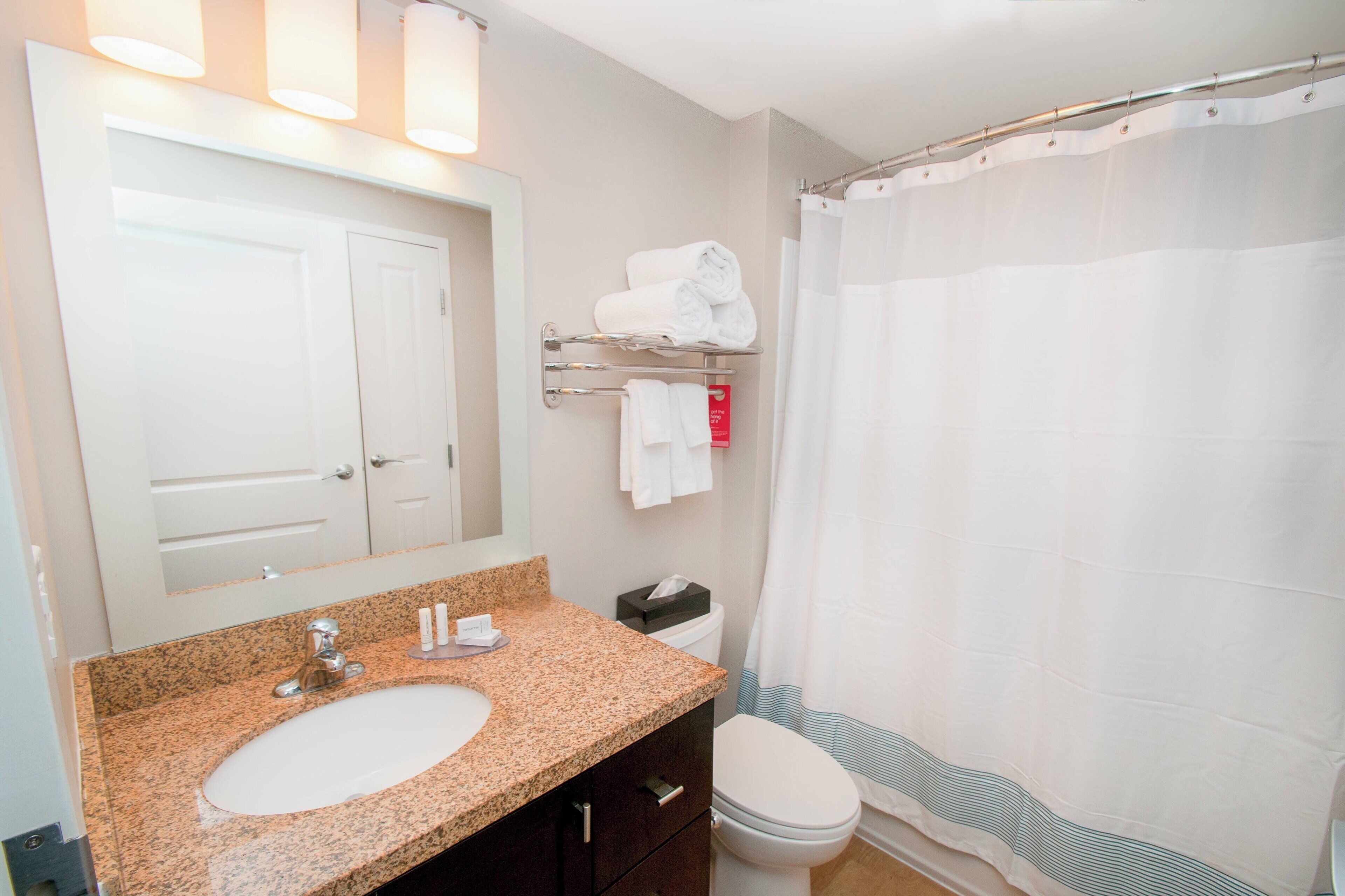 A bathroom with a granite countertop sink, a white toilet, a towel rack with folded towels, and a shower curtain.