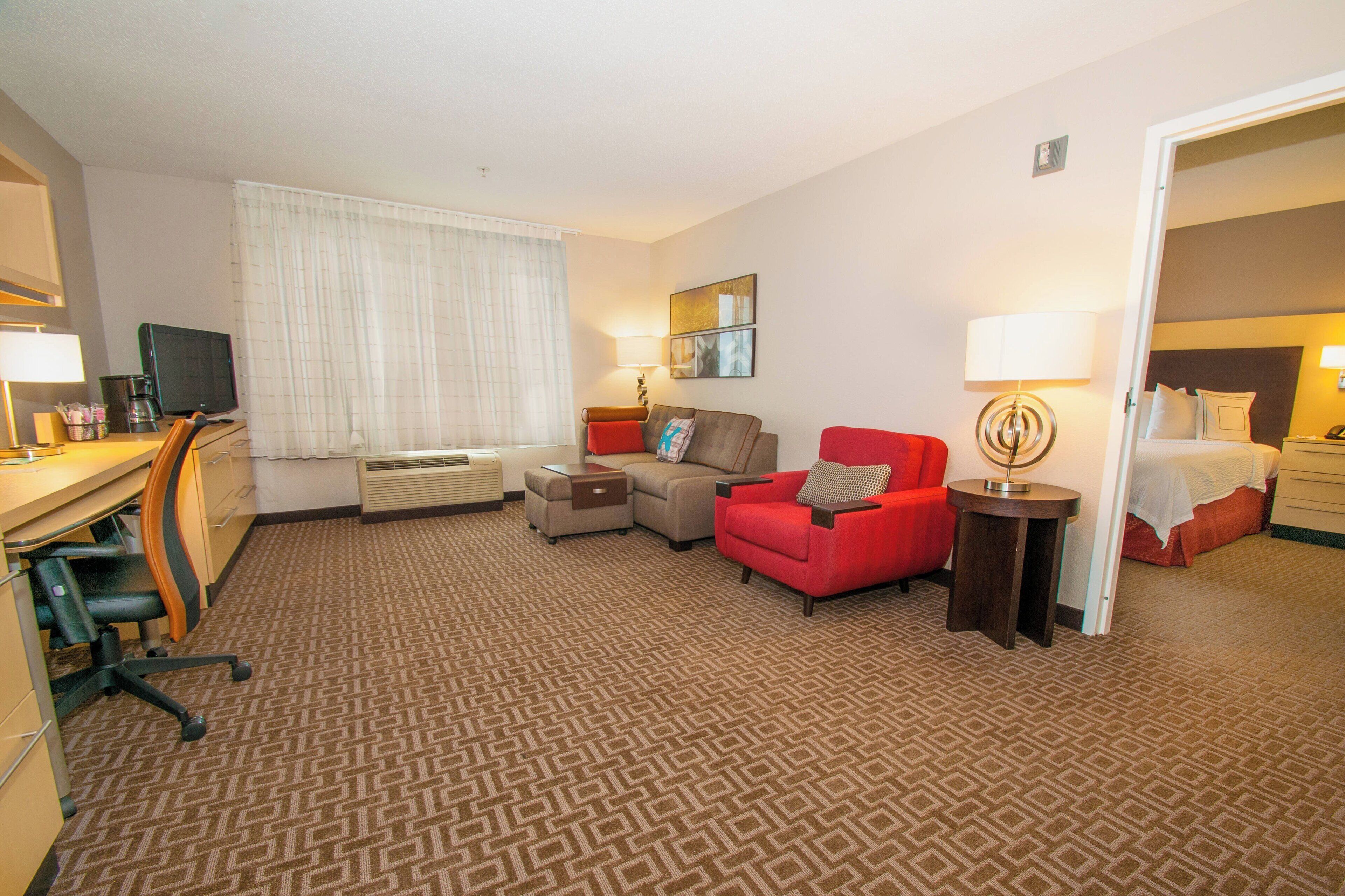 A hotel room with a desk, two chairs, a sofa, a bedside table with a lamp, a bed, and a window with curtains.