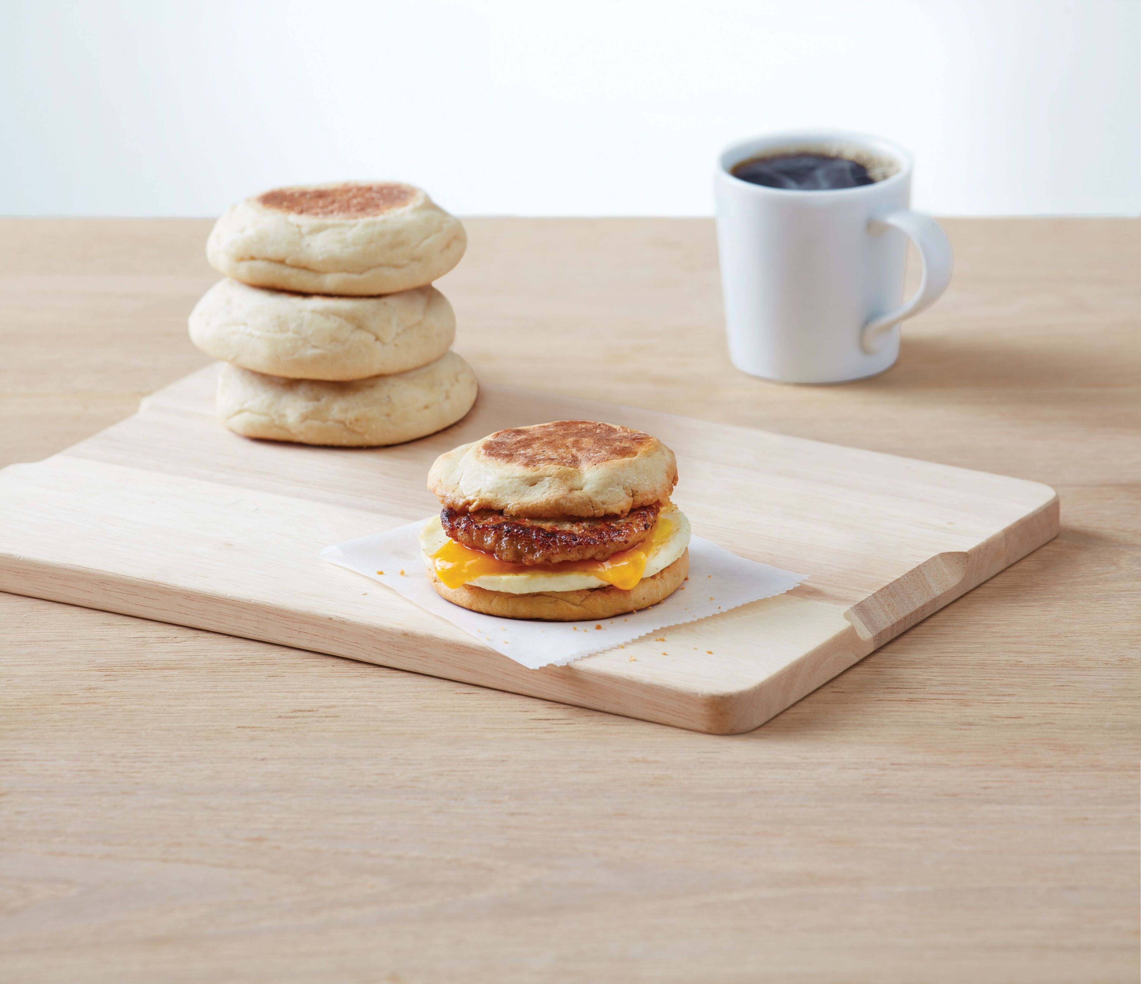 A breakfast sandwich on a wooden board with a stack of English muffins and a white mug in the background.