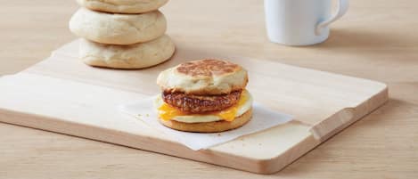 A breakfast sandwich on a wooden board with a stack of English muffins and a white mug in the background.