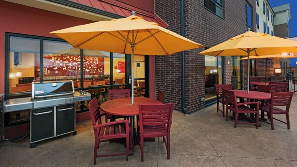 Een buitenterras met rode tafels en gele parasols, een barbecuehoek, en een bakstenen gebouw op de achtergrond.