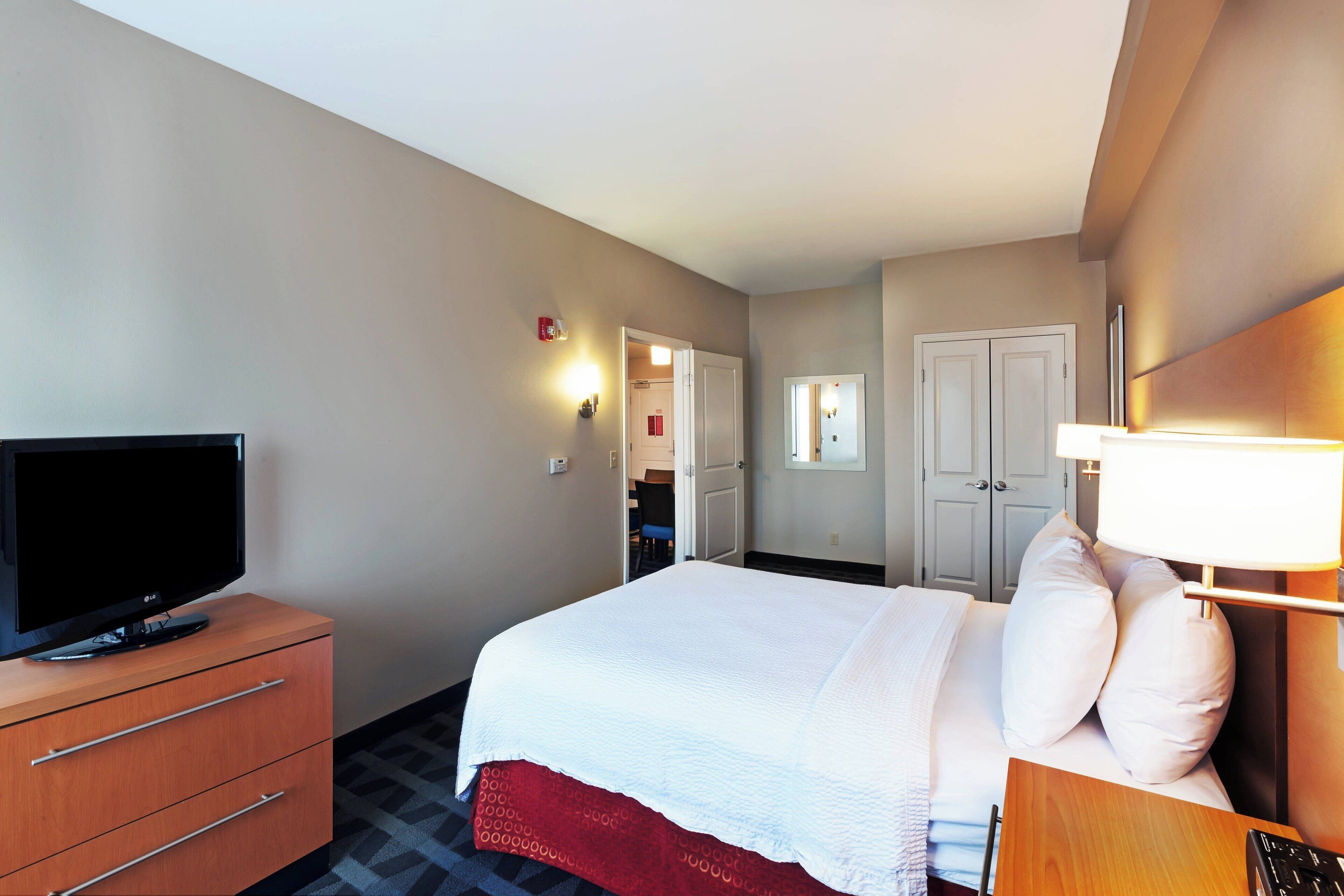 A hotel room with a bed, a television on a wooden dresser, a desk with a lamp, and a door leading to a bathroom.