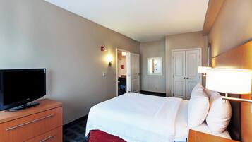 A hotel room with a bed, a television on a wooden dresser, a desk with a lamp, and a door leading to a bathroom.