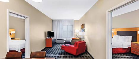 A hotel room with a bed, a red armchair, a wooden table, a television, and a desk with a lamp.