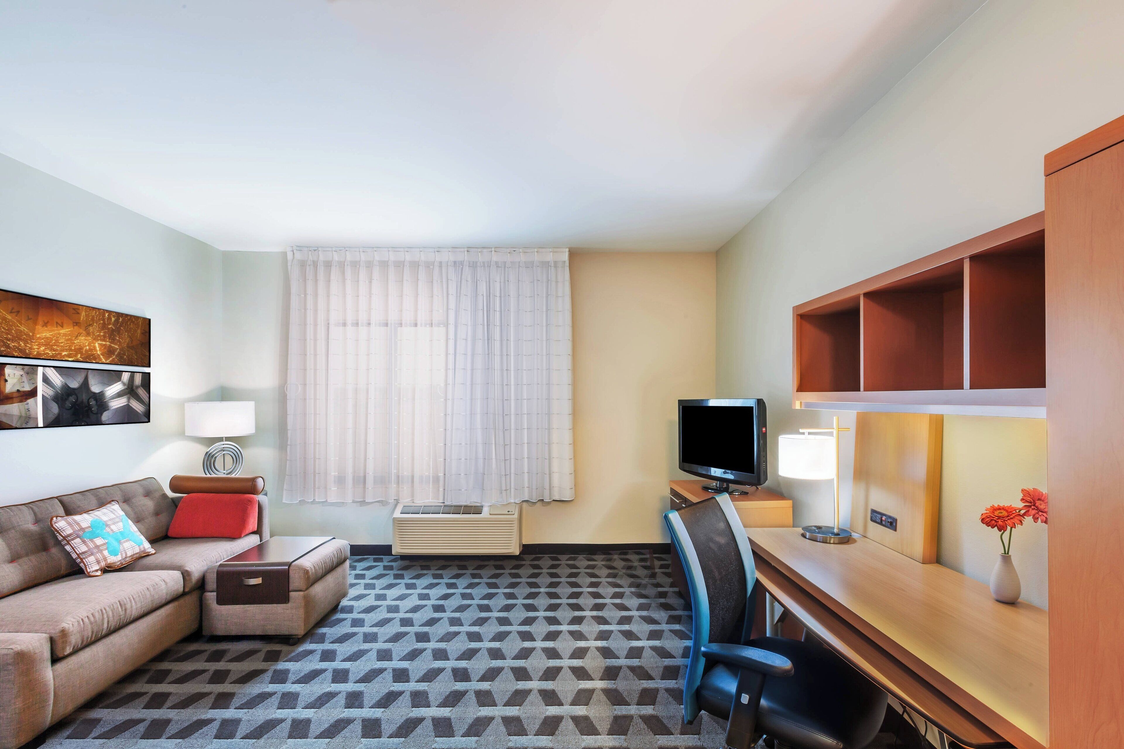 Habitación de hotel con sofá, escritorio, televisión, lámpara, ventana con cortinas y alfombra estampada.