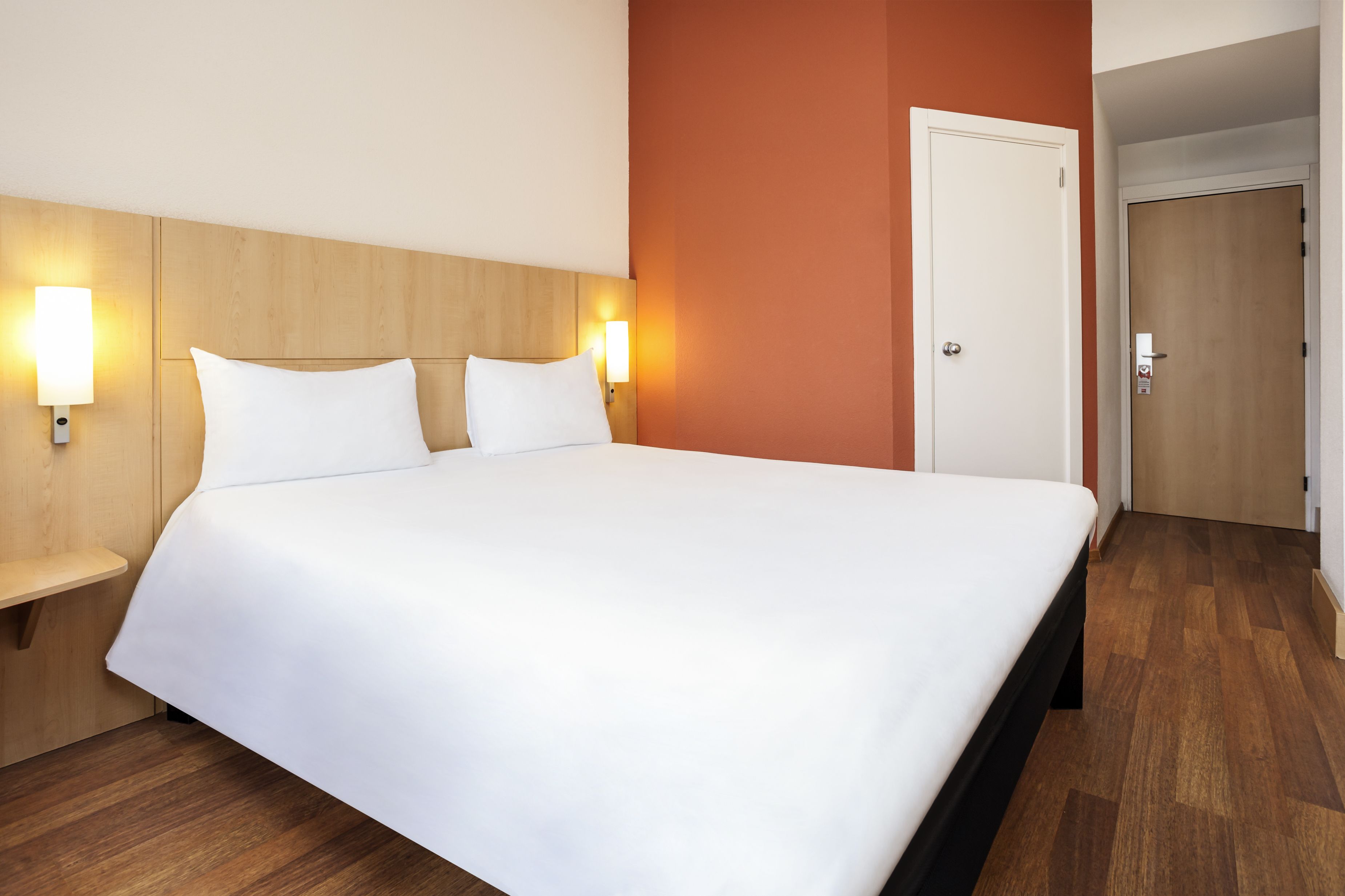 Een hotelkamer met een groot bed, een houten hoofdbord, twee bedlampen en een deur naar een andere kamer.