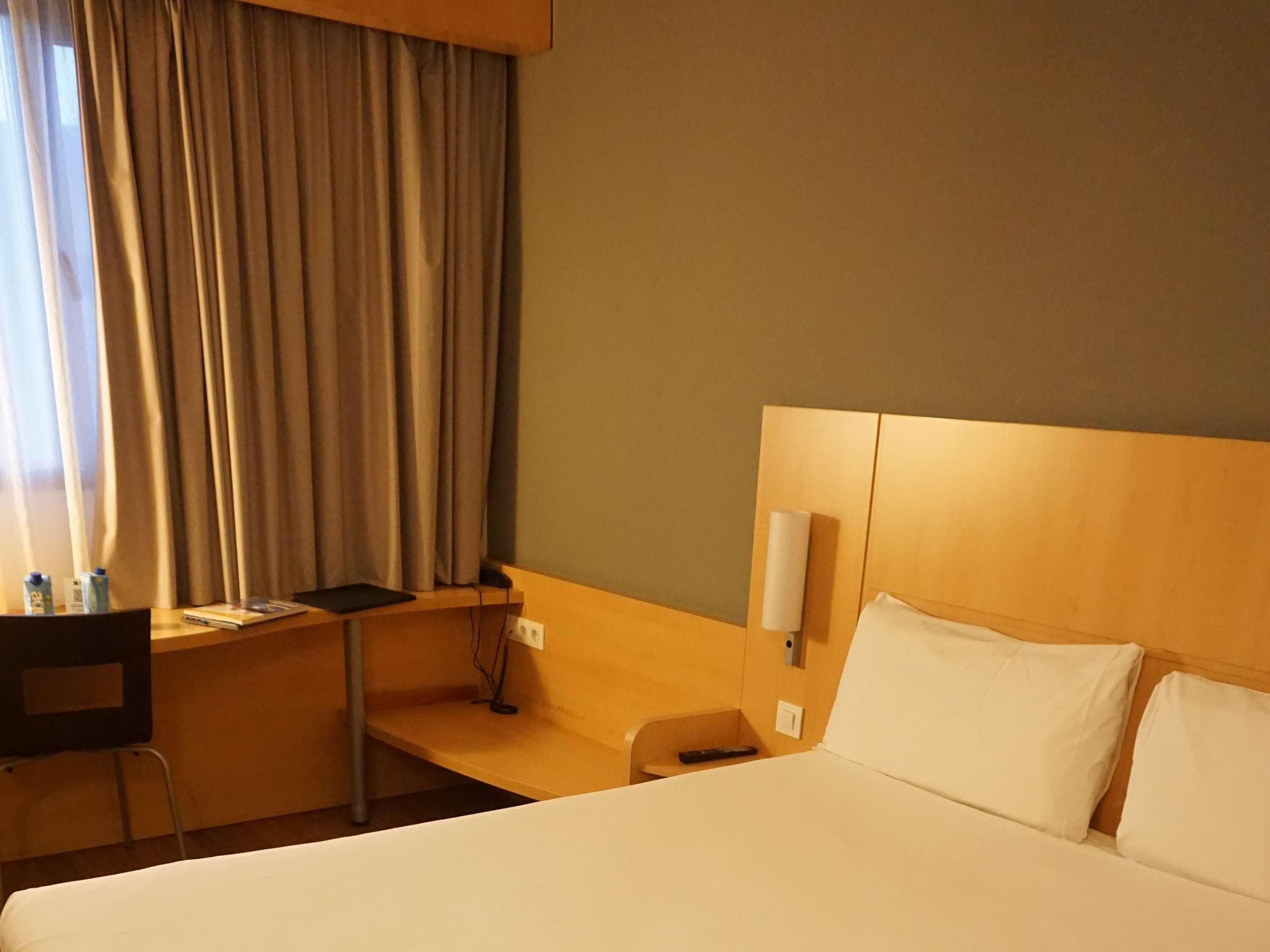 Habitació d'hotel amb llit, escriptori, cadira, làmpada i finestra amb cortines.