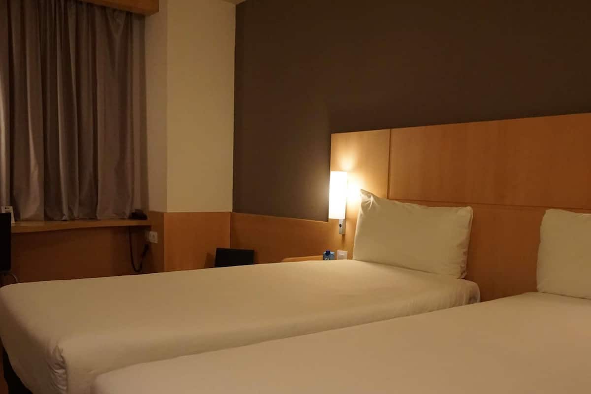 A hotel room with two beds, a nightstand, a lamp, and a window with curtains.