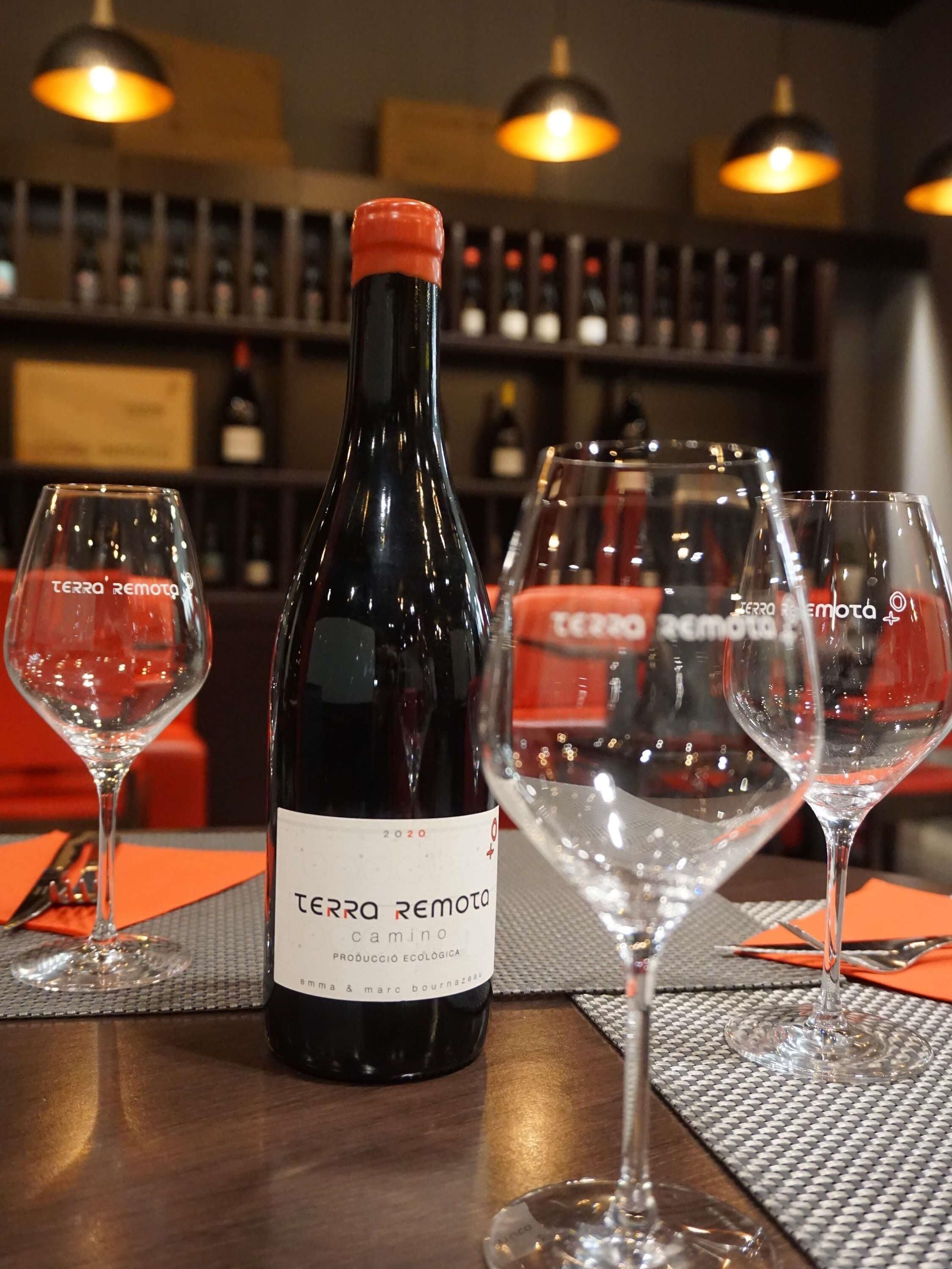 A bottle of Terra Remota wine with two glasses on a table, with a bar and shelves of bottles in the background.