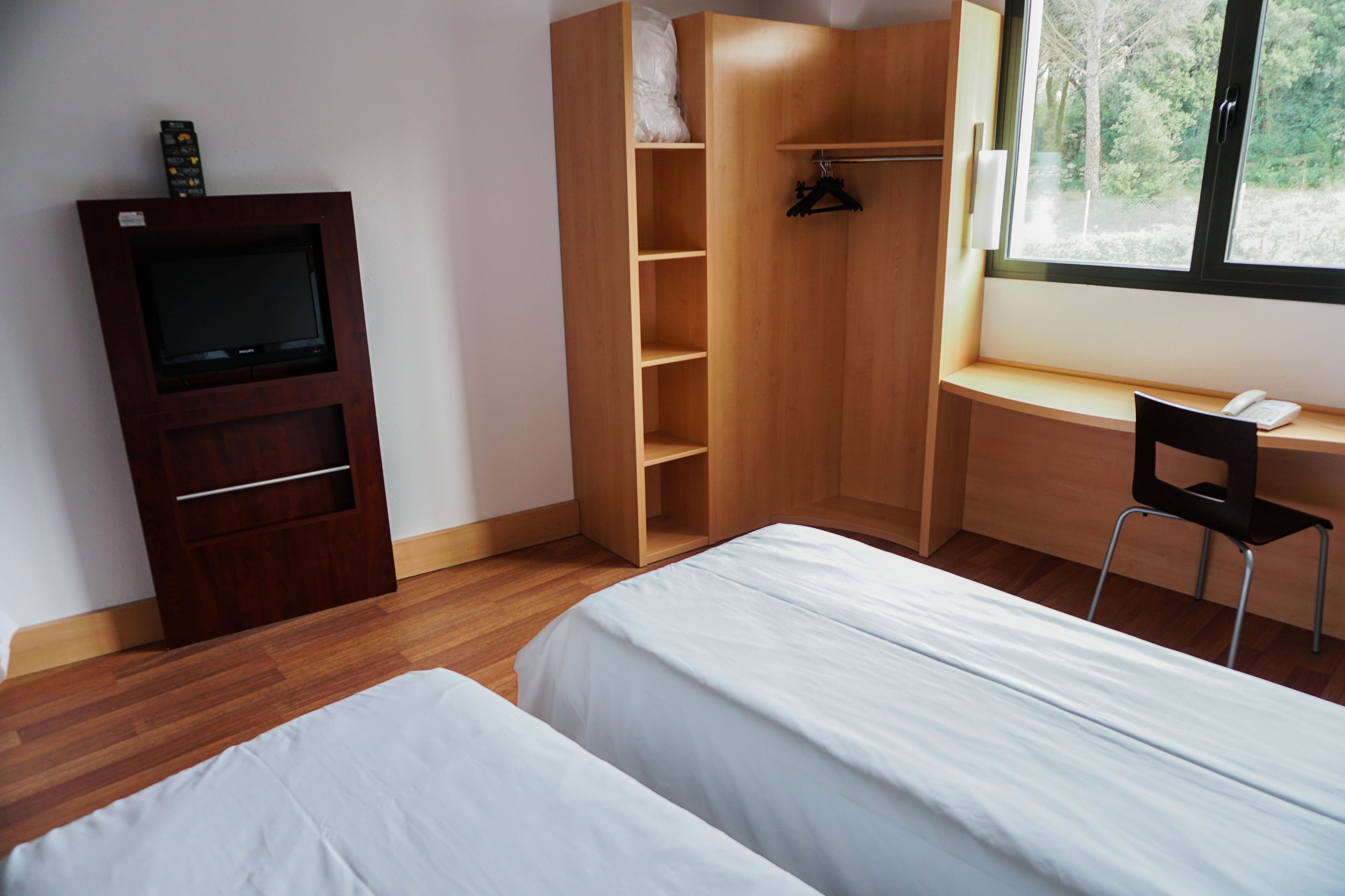 A bedroom with two beds, a wooden wardrobe, a TV, and a desk with a chair.