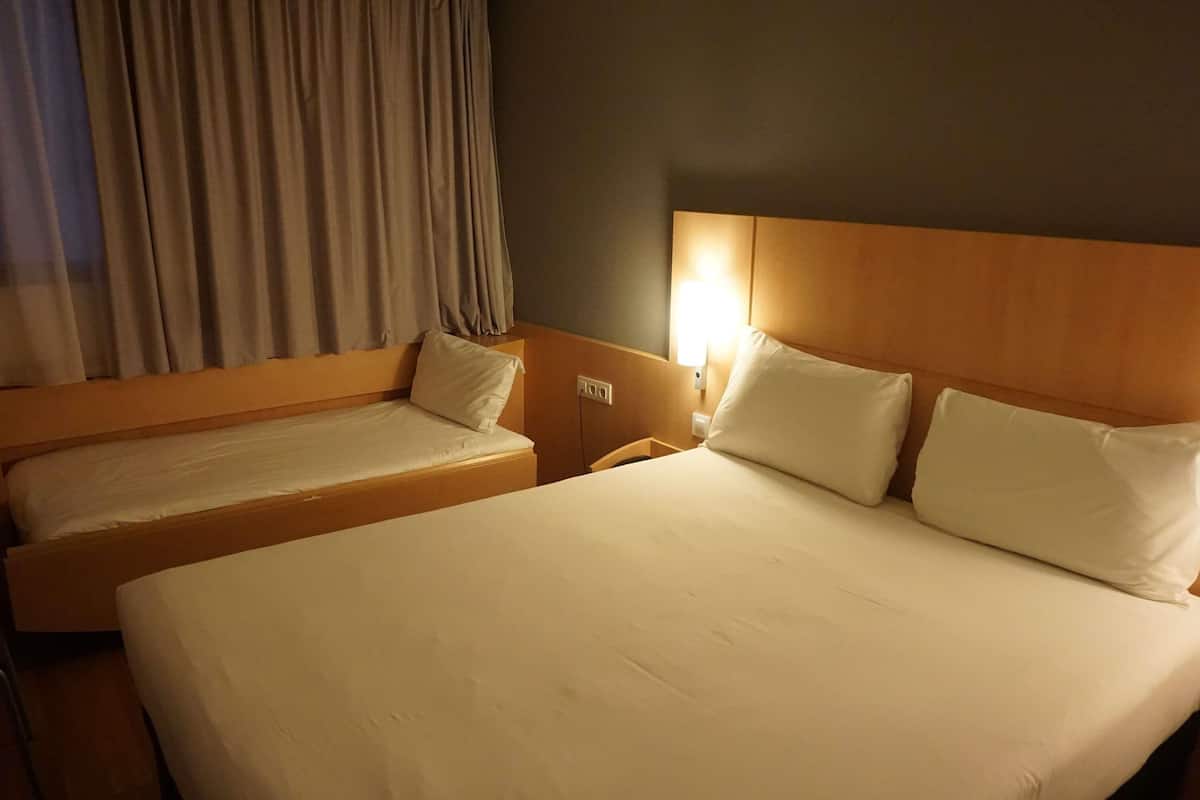 A hotel room with two beds, a nightstand, a lamp, and a window with curtains.