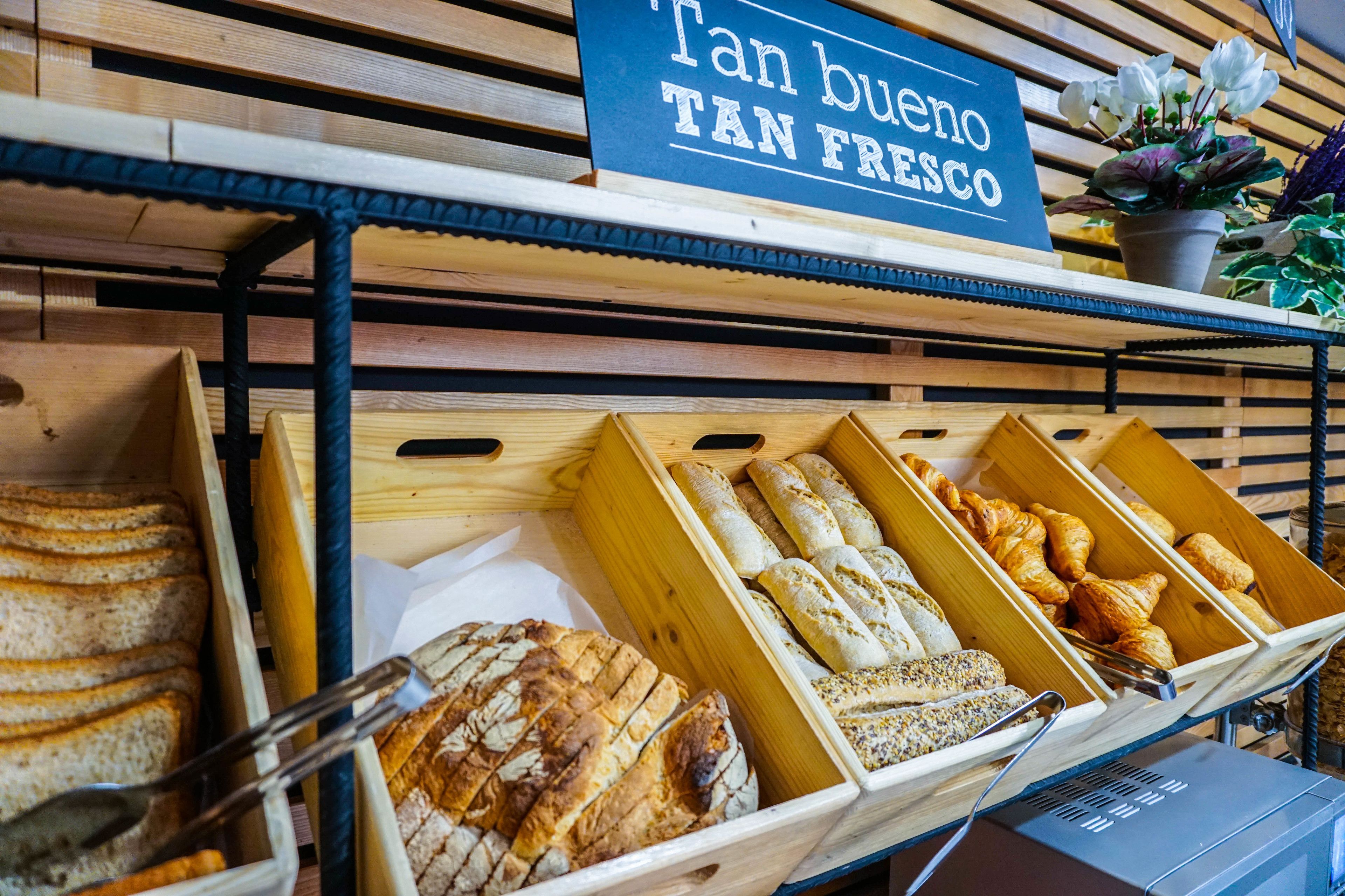 Variedad de panes y pasteles exhibidos en cajas de madera, con un letrero que dice "Tan bueno TAN FRESCO".