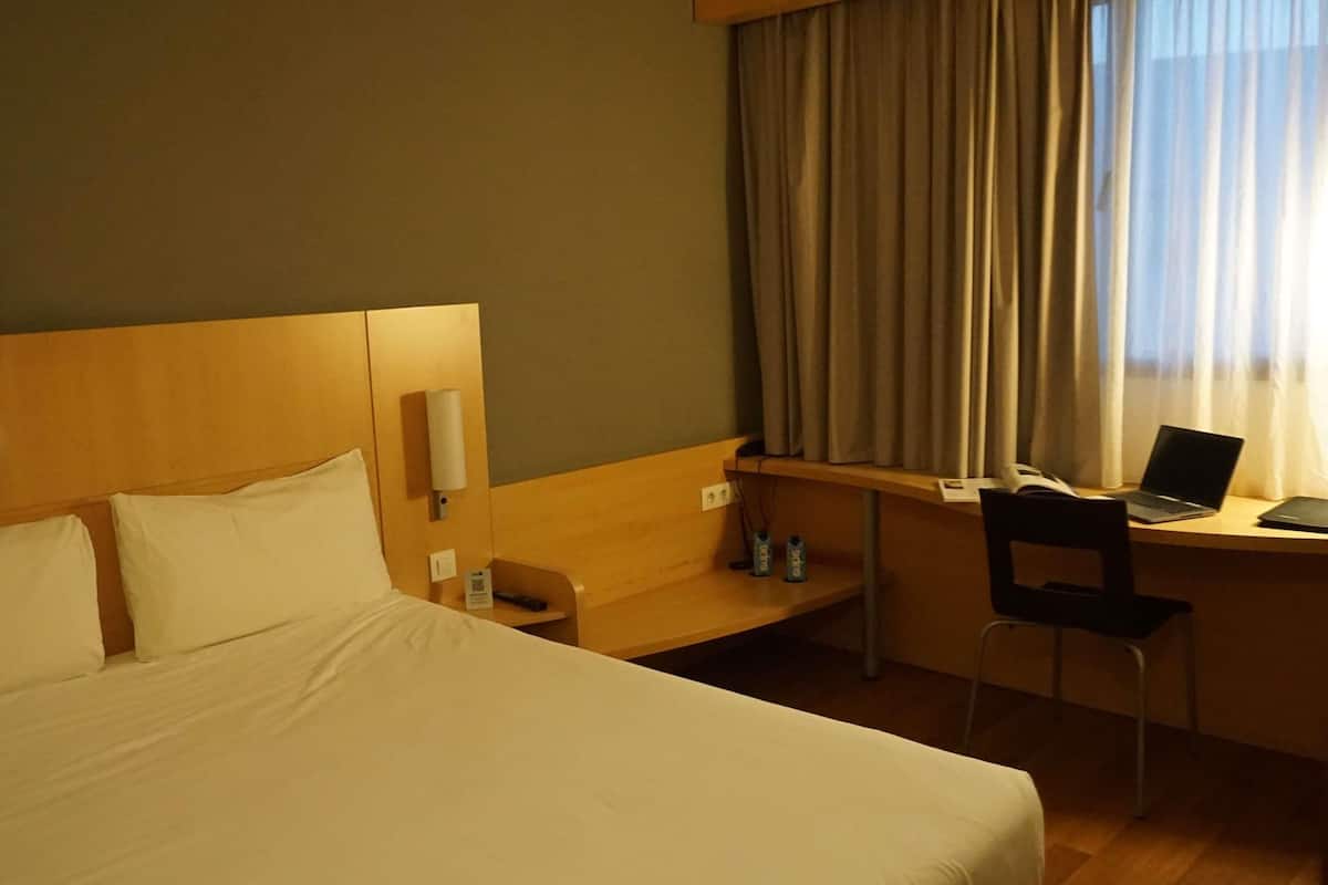 Una habitación de hotel con cama, escritorio, laptop, silla y ventana con cortinas.