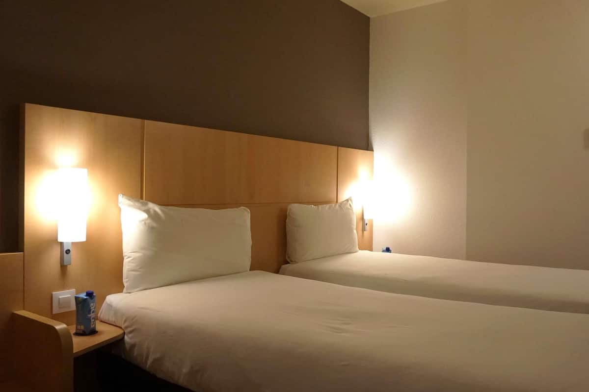A hotel room with two beds, a wooden headboard, bedside lamps, and a nightstand with a water bottle.