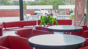 Un restaurante con asientos rojos y mesas redondas, que ofrece vistas a un vehículo estacionado y un edificio al fondo.