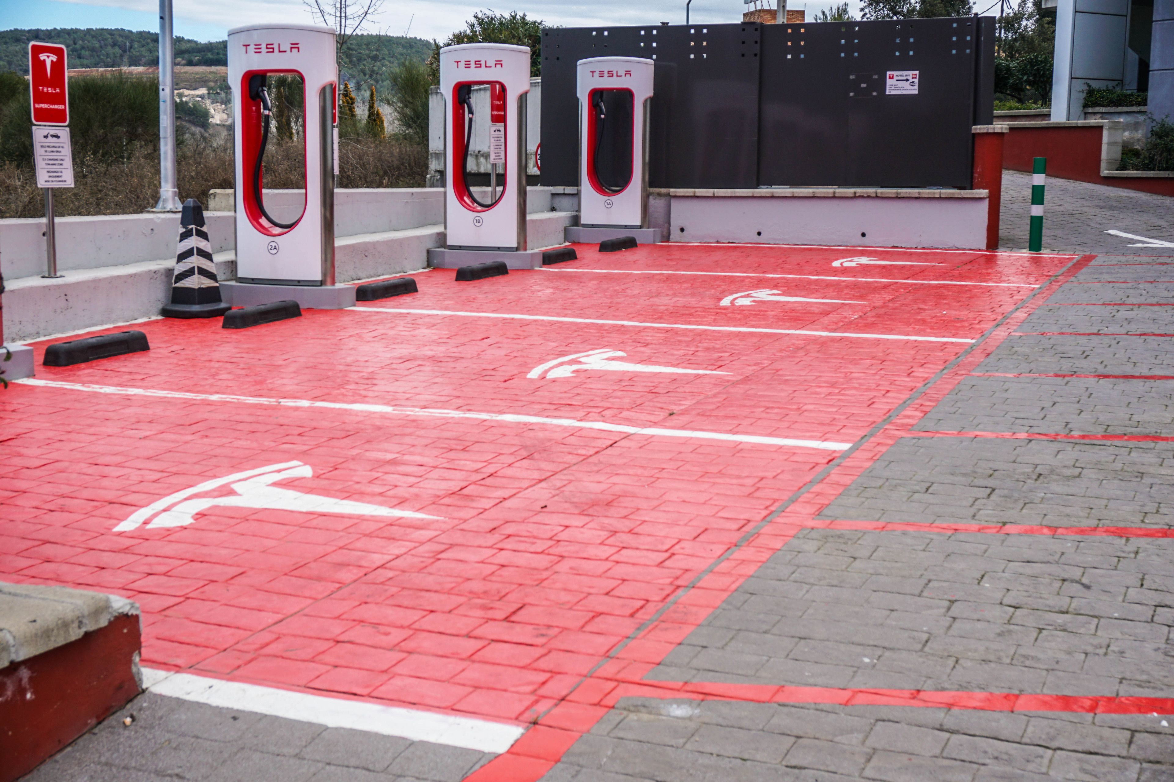 Una estación de carga Tesla con tres puntos de carga, un pavimento rojo con flechas direccionales blancas y una valla negra al fondo.